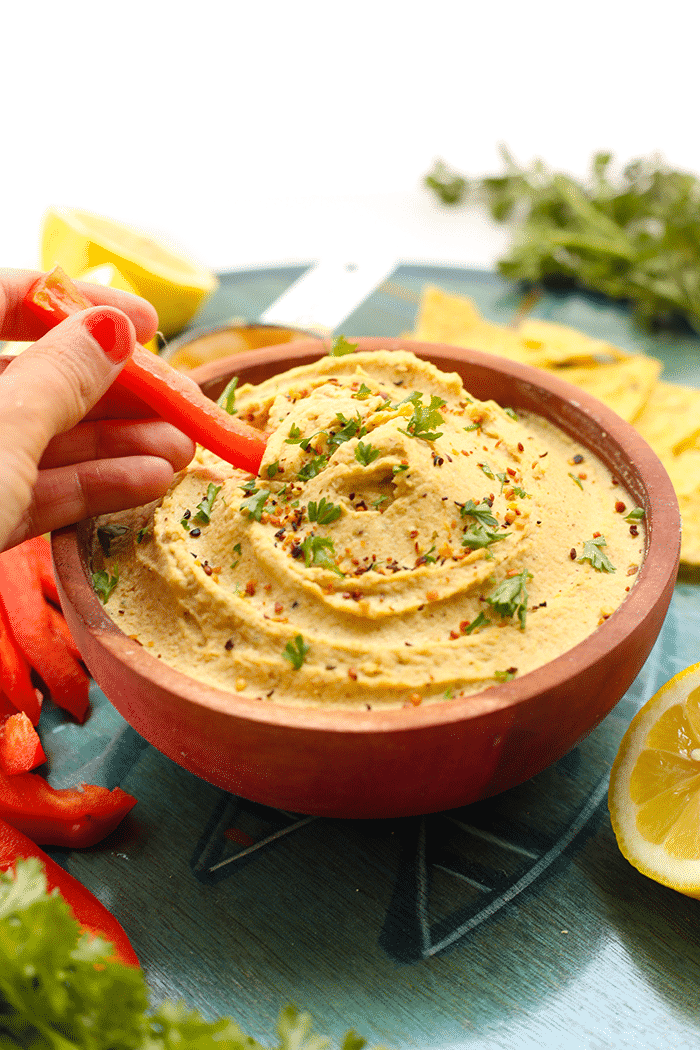 Bowl of lemon hemp hummus with a hand using a slice of red bell pepper to scoop some hummus out.