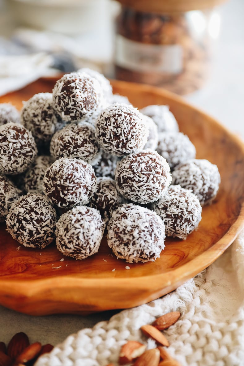 Almond joy energy balls stacked on a wooden platter. Cream towel with almonds scattered in the foreground.