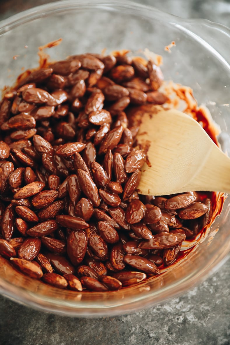 Almonds in a clear mixing bowl getting coated with melted chocolate by a wooden spoon.