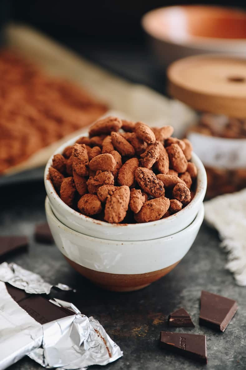 Angled shot of dark chocolate almonds in a white ceramic bowl. Chocolate pieces in the foreground.