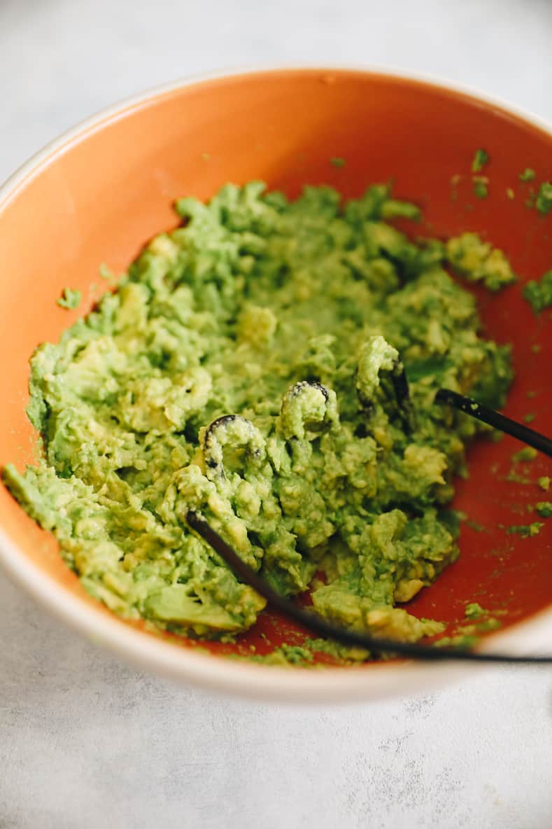 Avocados mashed in orange mixing bowl.