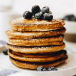 Stack of whole wheat pancakes on a white plate topped with fresh blueberries and a sprinkle of powdered sugar.