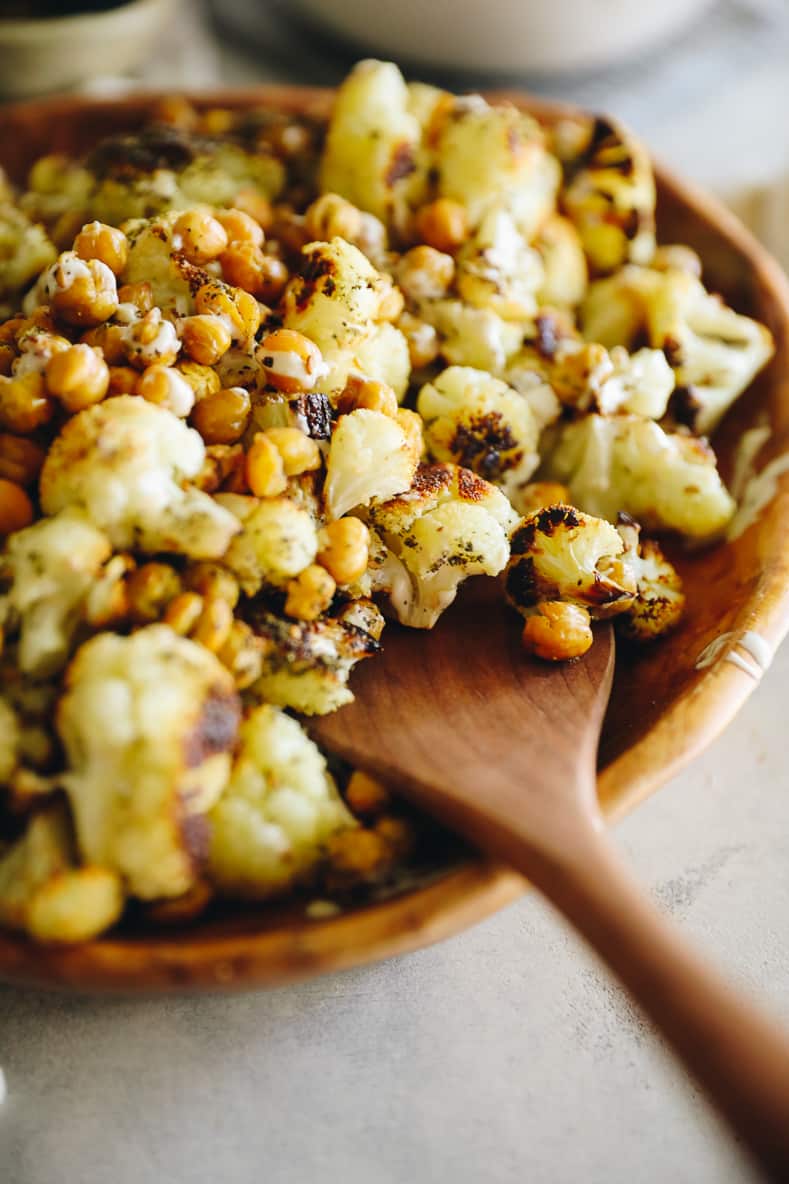 A wooden serving bowl filled with roasted cauliflower and chickpeas. Wooden spoon sitting on the edge of the bowl.