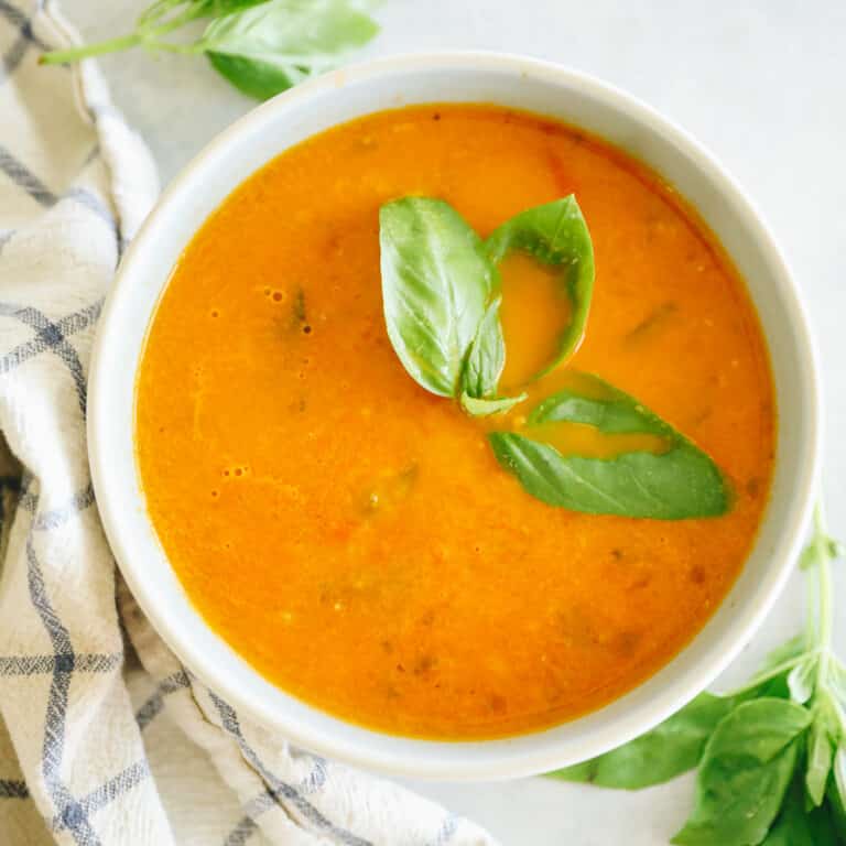 Overhead shot of roasted tomato basil soup in a white bowl. Basil leave garnish the soup and surround the bowl.