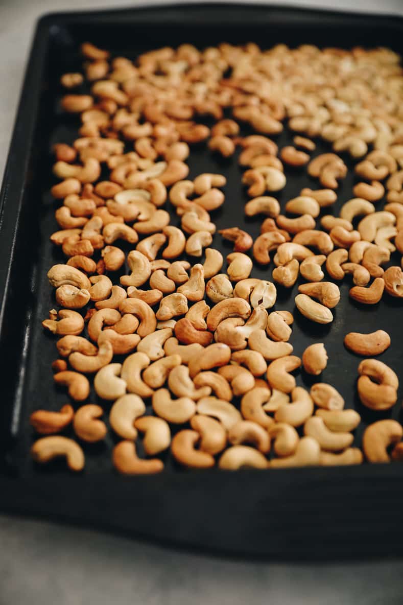 Cashews spread out on a black baking sheet.