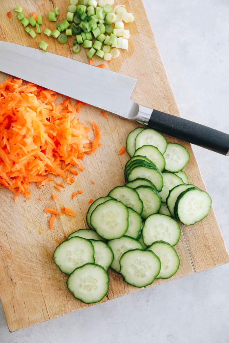 A cutting board with sliced cucumbers, shredded carrots and chopped green onions.