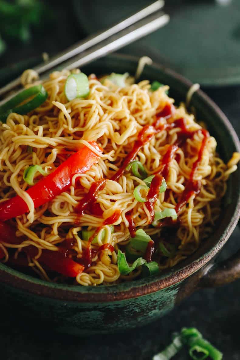 close-up of spicy ramen noodles in a green bowl with chopsticks and a drizzle of sriracha sauce.