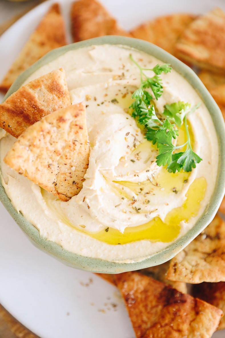 overhead image of homemade hummus in a green bowl with pita chips, olive oil, za'atar and fresh parsley