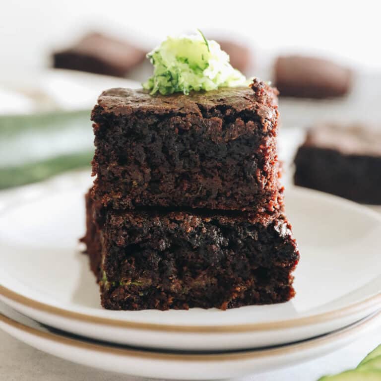 zucchini brownies stacked on a white plate with grated zucchini on top.